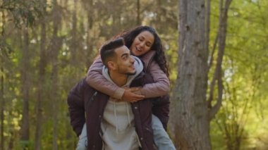 Happy young carefree married couple in love on romantic date in park in nature joyful girl sits on piggyback hugging man by shoulders enjoys freedom with arms outstretched to side talking laughing