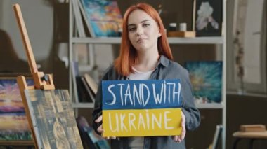 Red-haired talented artist stands posing in art studio workshop against background of drawing picture, painter holds cardboard banner stand with Ukraine support freedom of Ukrainians anti war poster