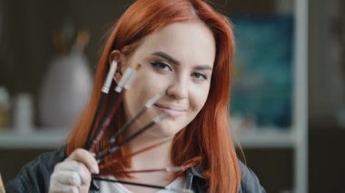 Kızıl saçlı genç kadın resim öğretmeni ressam kız ressam elinde fırça tutarken poz verirken kameraya gülümsüyor. Resim öğrencilerinin resimlerini çizdikten sonra yaratıcı stüdyo sahibi