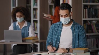 Kütüphane öğrencileri koruyucu maskeli genç Hintli adam masaya oturup sınav için ödev hazırlıyor notlar yazıyor mutlu adam kameraya bakıyor ve işaret işareti yapıyor.