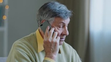 Caucasian old grey-haired man grandfather at home talking on mobile phone remote conversation distant consultation with doctor answering call emotionally speaking with smartphone connection service