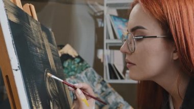 Yan görünüş, kızıl saçlı, 20 'li yaşlarda, gözlüklü, yetenekli, kadın ressam, siyah zemin üzerine resim çiziyor. Altın yağlı akrilik kadın, tuvale resim çiziyor. Sanat eğitimi alıyor.