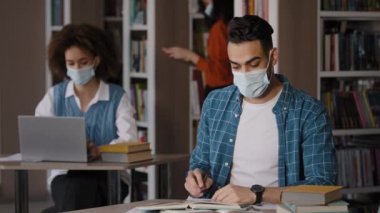 Koruyucu maskeli genç bir öğrenci sınav için notlar yazar deneme yazıları yazar tamam işaret işareti gösterileri kütüphanede yüksek öğrenim için yüksek öğrenim önerir.