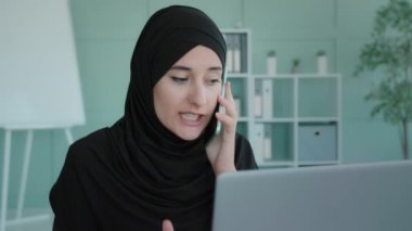Angry Islamic Muslim Arabic woman working with laptop shout scream speaking on mobile phone in office discussing problems conflict business trouble at work negative emotions conversation bad calling