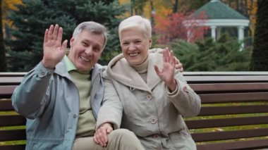 Caucasian married couple family retired 60s people man woman waving hands hello goodbye gesture elderly grandparents sit on bench in autumn park lovely talking gesticulate having pleasant conversation