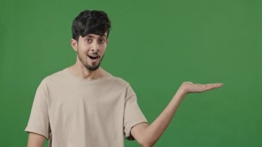 Indian man smiling recommend blank area over green background holds copy space on hand advertise points at empty place arabic guy using mockup for promo advertisement thumb up finger best choice sign