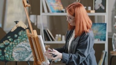 Yandan bakan sanat işçisi, gözlüklü, kızıl saçlı, yetenekli sanatçı, gözlüklü, resim sınıfında çalışan, boyalı, motivasyonlu ressam, boya fırçası yağlı akrilik resimli, soyut bir resim çiziyor.