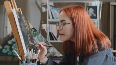 İlham verici, odaklanmış, kızıl saçlı bir kadın ressam gözlük takıyor. Ağızda fırçalar, tuvalde yağlı boya boya boya, stüdyoda ilham verici güzel çizimler yapıyor.