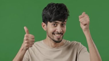 Friendly arabic man stand in green studio make thumb up gesture both hands show like everything okay smiling young hispanic guy looking at camera wining recommend best choice successful agreement sign