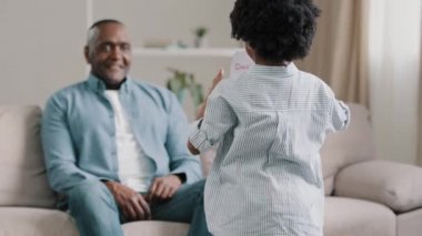 African american man sitting on sofa in room accepting congratulations caring daughter gives card to beloved dad on fathers day cute kid girl wishes happy birthday male surprised by unexpected gift