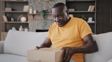 Happy surprised african senior man open cardboard box with gift unpacking parcel delivery internet purchase american mature adult homeowner sitting at home sorting keys pen postcard from open package