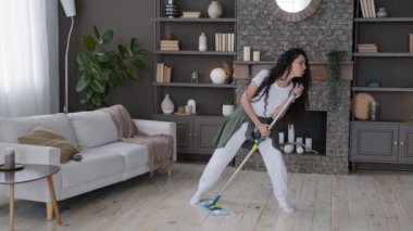 Funny young hispanic woman housewife cleaning at home washes floor in living room cheerful excited girl expressively sings uses mop as microphone performs movement energetic shaking long curly hair