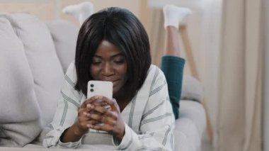 Happy beautiful african american girl lying on cozy sofa resting holding phone young woman chatting with friends online browsing web page making order in internet using smartphone checking dating site