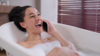 Joyful young woman lies in foam bath relaxes in luxurious bathroom satisfied attractive girl speaks on phone answers friendly call laughs chatting with friend uses smartphone for remote communication