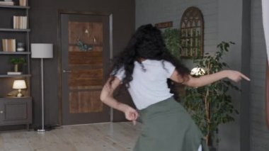 Arkadan genç heyecanlı bir kadının modern oturma odasında dinamik dans edişine bakın enerjik kız blogcu dans içeriği içeriği vlog duyarlı kadın aktif hareket yapıyor hafta sonu kanepeye düşüyor ve eğleniyor.