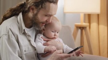 Kafkasyalı baba sakallı, yeni doğmuş bir kızı olan bir çocuğu evde tutuyor. Cep telefonuyla online akıllı telefon alışverişi videosu çekiyor. Yüksek kalite 4k görüntü