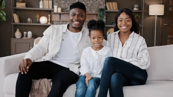 African family sit down on comfortable couch in living room. Biracial parents and child smiling looking at camera. Cheerful daughter with mum and dad at home sitting on sofa in new house, real estate