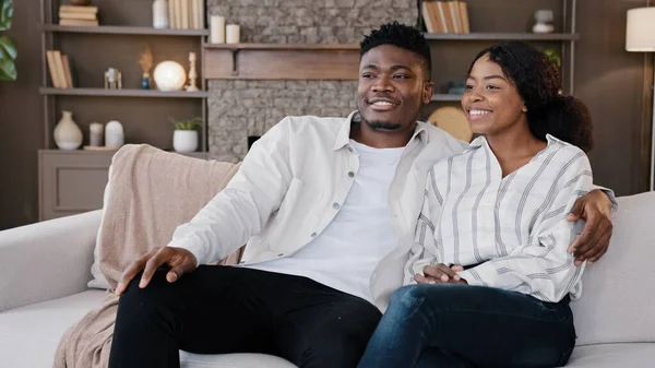 Happy newlywed couple married African man and woman sitting at home sofa watching TV talking laughing. Husband and wife talk with family therapist. Boyfriend and girlfriend casual conversation at home