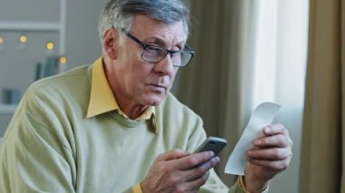Kafkasyalı 60 'lı yaşlarda yaşlı bir adam akıllı telefon tutuyor cep telefonu kullanıyor e-banka uygulaması ile muhasebe faturalarını hesaplıyor kağıttan çek ile telefonda hesap makinesi uygulaması kullanıyor