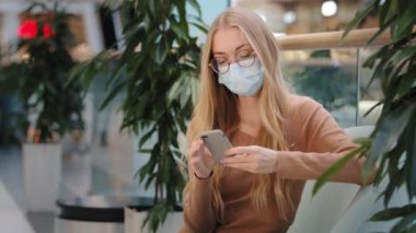 Caucasian blonde woman wearing medical mask in shopping center mall company looking at phone screen chatting online free Wi-Fi cellular connection 5g girl female sitting on couch using mobile text app
