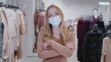 Successful blonde young woman seller consultant girl clothing store owner small business lady buyer customer client in medical mask standing in mall looking camera posing with arms crossed over chest