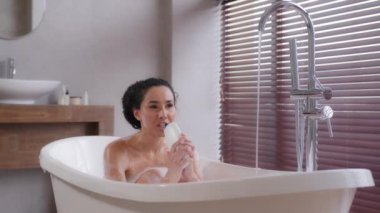 Funny young woman sits in foamy bath sings using shampoo bottle as microphone enjoying energetic morning daily hygiene routine carefree happy emotional girl singing song in bathroom feels joy pleasure
