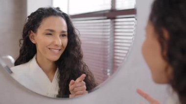 Pretty confident young woman stands in bathroom looks in mirror winks at reflection motivating for success attractive girl enjoys natural beauty after beauty treatment morning daily routine concept