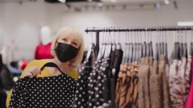 Mature caucasian female sales assistant in protective mask dances in clothing store advertises new stylish collection of clothes with discount happy woman buyer finds right date outfit enjoys purchase