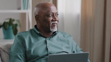 Focused old senior bald overweight African man freelancer mature male working on computer at home sitting on couch looking in distance searching inspiration thinking about idea creating online project