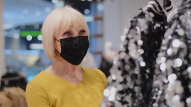 Giysi mağazasında koruyucu maske takan olgun bir kadın, parlak kıyafete hayran kalır. Yaşlı bir bayan şaşaalı elbiseye şaşırır. Kaliteyi jest onayı ile değerlendirir.
