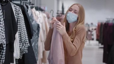 Blonde caucasian girl woman shopper in medical mask chooses perfect casual blouse finds clothes dancing hugging cuddling jacket rejoices in black friday shopping discounts happy buying in store. High