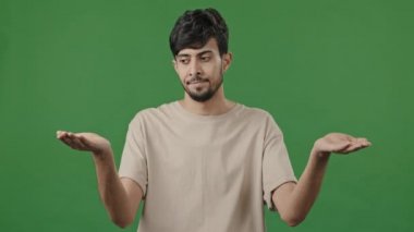 Puzzled man confused uncertain hispanic arab thoughtful young guy standing in green studio shrugs shoulders make choice decision choosing doing weights gesture with hands compares between two options