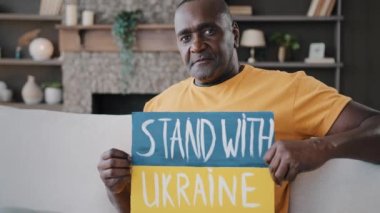 Ciddi, yaşlı, Afrikalı, yetişkin, 50 'li yaşlarda, bekar, bekar bir erkek, evinde, koltukta oturuyor. Mavi renkli, sarı renkli bir karton. Üzerinde Ukrayna destekleyici sembolü olan harf işareti var.