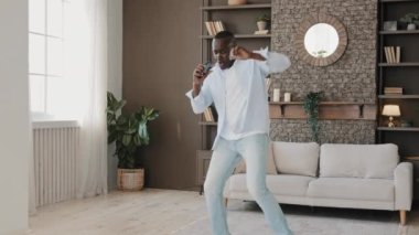 Senior african american comical adult man active moving dancing in modern living room singing loud favorite song in phone use mobile as microphone enjoying leisure at home alone rhythmic dynamic dance