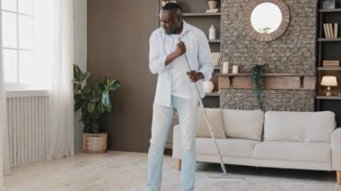 Excited artistic adult african man householder bachelor active washing floor cleaning parquet with mop at home living room moving to music pretending solo singer using housekeeping swab as microphone