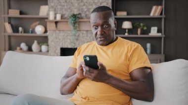 African american elderly man winner user senior grandpa biracial successful consumer buyer sit at home on sofa hold phone read news scroll website pages play mobile game win online lottery bet victory