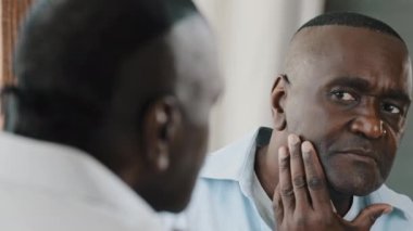 Close up male reflection in mirror african american senior bristly man touching stubble face dissatisfied with wrinkles doing morning beauty routine stare looking at himself preparing for working day