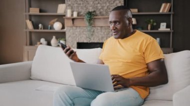 Odaklanmış kıdemli Afro-Amerikan erkek alışverişçi müşteri ev işi yapıyor yaşlı iş adamı serbest çalışıyor dizüstü bilgisayara bak internetten alışveriş rezervasyonu yap cep telefonu konuş istemci