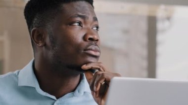 Thoughtful african american businessman writer dreamer genius man biracial guy holds hand on chin creates business plan come up with idea for startup uses computer typing text creates internet project