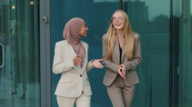 Diverse businesswomen colleagues walking outdoors communicate informally, discuss business meeting or deal. Indian girl in hijab laughing with colleague young European woman during office break . High