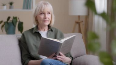 Calm smart Caucasian 50s lady dreaming old woman mature lady senior grandmother professor teacher sitting on sofa resting studying education relaxing at home reading book enjoying literature learning