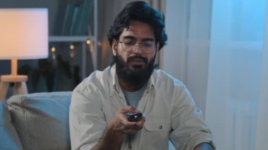 Hispanic Indian middle-aged adult bearded man in glasses sits on sofa at home in evening at night switches channel watching TV movie online broadcast of football match enjoying cinema film series
