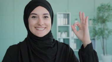 Portrait indoors Muslim Islamic girl smiles toothy wide at camera showing hand ok sign. Female Arabian student shows everything fine success gesture body language good idea agree advertising gesture