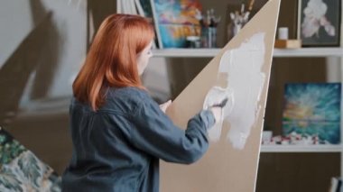 View from behind red-haired Caucasian girl student creative woman artist painter designer scenery picture on canvas cardboard plywood smears white plaster with spatula preparation painting art design