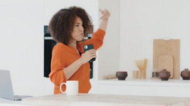 Active cheerful funny girl in orange blouse with African hairstyle with curls biracial women housewife at home kitchen dancing having fun singing into mobile phone like microphone listening to music
