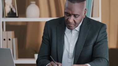 African American businessman mature man boss leader manager banker professor 60s worker sits in office table writes notes information in document looking at laptop prepares for lecture analyzes data