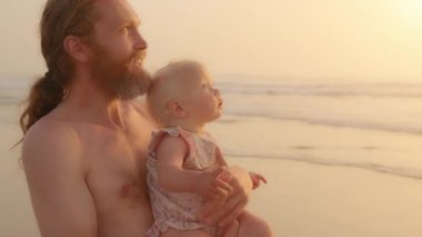 Caucasian bearded father holding little baby infant small daughter son newborn standing at sandy beach near sea ocean watching in gold sky dad talking explain showing direction with finger pointing