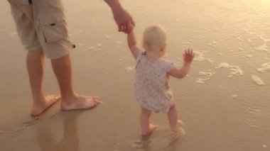 Unrecognizable Caucasian caring father holding baby daughter toddler by hand stepping walking on wet sand barefoot playing with waves in sea ocean on beach enjoying hot day on vacation explore nature