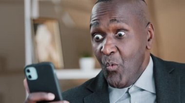 African American old 50s surprised amazed shocked man looking at mobile phone screen reading unbelievable news waving head no disagreement negation refusal shock fear amazing online sms shock reaction