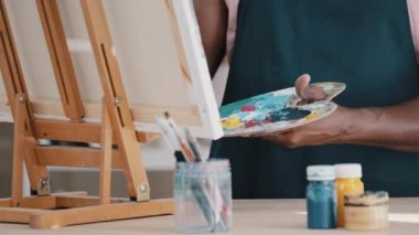 Close-up male hands holding palette mixes acrylic paints painting picture. Unrecognizable African man artist drawing with paint brush in studio art therapy unknown painter in apron create masterpiece